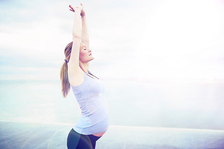 Attractive young pregnant woman doing breathing exercises at the seaside standing with her arms raised above her head and eyes closed in a fitness and health concept